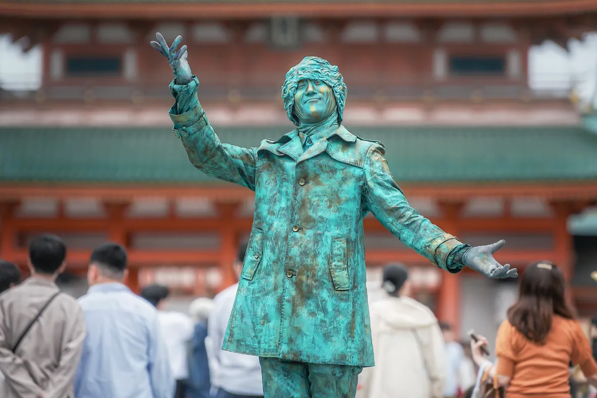 朱塗りの門を背に、片手を高く挙げ、もう一方の手を広げて立つ緑青色の石像姿の Mr.Kids。手前には通行人の後ろ姿